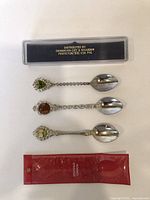 Photo showing three souvenir spoons with decorative handle emblems and two display cases, one clear black-backed case and one red pouch, displayed from top view.