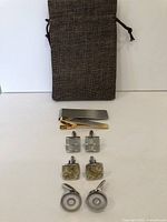 Two tie clips and two money clips with four pairs of cuff links displayed in front of a brown drawstring jewelry pouch.