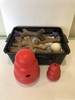 Black basket filled with seven brown bone-shaped rubber chew toys, a white ball, and a purple knotted rope toy, with a folded white and blue striped blanket under them. Two red Kong rubber toys placed in front of basket on white surface.