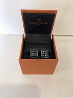 Front view of square Simon Carter cuff links with black enamel and silver grid pattern displayed inside black cushioned interior of orange presentation box.