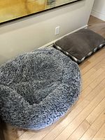 Two dog beds placed on a hardwood floor next to a beige sofa and wall outlet. One is a round grey fluffy bed and the other is a rectangular brown cushioned bed with plaid sides.