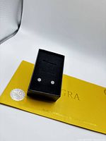 Pair of moissanite stud earrings set in black jewelry box placed atop a yellow folder labeled GRA, displaying the certificate of authenticity.