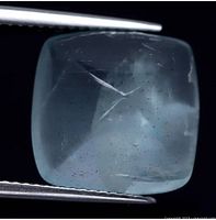 Close-up photo of a light blue, cushion-shaped aquamarine cabochon held by tweezers showing visible internal inclusions and polished smooth surface.