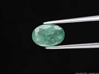Oval cut Zambian emerald gemstone held by tweezers, showing top view with medium green color and inclusions.