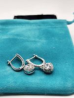 Pair of silver moissanite earrings displayed on a teal drawstring pouch. Earrings have round cut stones with secure hinged backs and small accent stones along the sides.