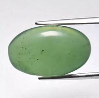 Close-up image of an oval cabochon green serpentine gemstone held by tweezers, showing polished smooth surface and natural inclusions.