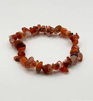 Close-up image of a strawberry quartz chip bracelet laid flat on a white surface, showing multiple tumbled stone chips in varied reddish-orange hues.