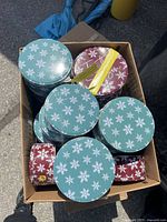 Photo of a cardboard box filled with over 20 round metal cookie tins with snowflake patterns in teal and maroon colors, two sizes small and medium.