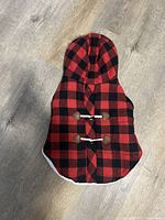 Front of red and black plaid dog coat with toggles and hood, laid flat on floor.