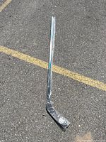 Full view of new black left-handed hockey stick wrapped in plastic on asphalt.
