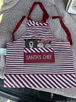 Photo of a kids Christmas apron with red and white stripes and a maroon patch featuring 'Santa's Chef' text and animal faces including a bear and a fox. The apron has red neck and waist ties.