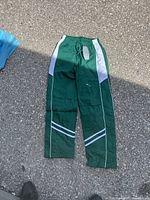 Green kids' sports pants laid flat on asphalt ground, showing white side stripes and elastic waistband with drawstring