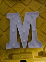 White light up letter M with bulbs placed on a yellow textured surface.
