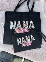 Photo showing a black fabric tote bag and matching wrist bag, both with 'NANA' printed in floral letters and pink flowers design.