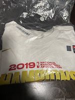 Folded white T-shirt with 2019 Champions gold and red print and barcode tag visible inside plastic bag.