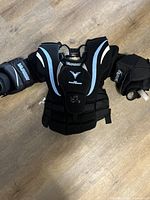 Full front view of the Vaughn hockey goalie chest protector laid flat on a wooden floor, showing the black and grey colors, padding, adjustable straps, and worn areas.