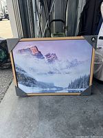 Framed wall art showing a mountain landscape with snowy peaks, frost-covered pine trees, and a still lake, set in a simple wooden frame with protective corner guards.