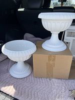 Two white urn planters of different sizes placed on a quilted blanket with one standing on a cardboard box and the other beside it.
