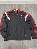 Front view of black Adidas AC Milan track jacket showing red three-stripe sleeves, white and red collar detail, embroidered AC Milan logo on left chest and Adidas logo on right chest.