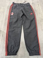Full view of black Adidas track pants with red three stripes, elastic waistband and cuffs, AC Milan crest and Adidas logo on front thighs.