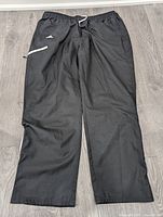 Full view of men's black Adidas casual track pants laid flat showing elastic waistband, white drawstring, side pockets, and Adidas logo detail.