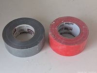 Two rolls of tape side by side on surface: silver/grey Nashua duct tape and red Ram Board Pro Tape.