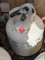 White empty propane tank placed on the ground outside with visible valve and safety stickers.