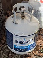 Photo of a white empty Bernzomatic propane tank featuring the top valve with protective guard, placed on outdoor ground among dead leaves.