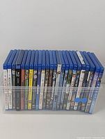 Photo showing a row of approximately 23 Blu-Ray movie cases standing upright in a clear plastic container. Cases are visibly mostly blue with multiple cover art designs on the spines.