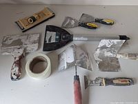 Overview of plaster and mudding tools including drywall hawk, mesh tape roll, multiple putty knives and scrapers with plaster residue