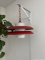 Red midcentury modern wall-mounted lamp with red metal layered shades and white inner glass shade, mounted on white wall bracket with visible electrical cord and nearby green plant.