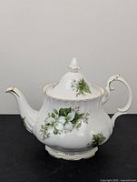 Front angled view of white bone china teapot with trillium flower pattern and gold trim on lid, spout, handle, and base.