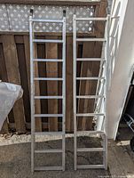Two aluminum trailer loading ramps leaning against a wooden fence, showing full length and ladder style steps.