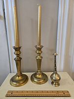Full view of two solid brass candle holders standing next to a smaller brass holy water bottle on a white surface, ruler in front for scale.