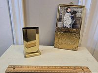 Both brass items displayed on a white surface with a measuring ruler in front for scale, showing the letter holder with framed image and eyeglass holder alongside it.