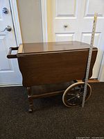 Antique wooden bar cart with fold-down side leaves closed, measuring tape showing approximate height, wooden handle and large wooden wheels visible.
