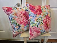 Two bright floral patterned square pillows with large pink flowers and multicolor accents, placed against a white door and on a small table.