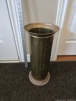 Front view of the tall brass umbrella stand with flared top rim and textured cylindrical body on floor near door.