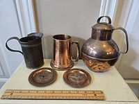View of four copper items and two small decorative copper plates with a ruler for scale on surface against a wall.