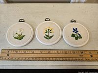 Three small round crewel floral wall hangings laid side-by-side on a flat surface with a ruler placed below for size reference, approximately 3.5 inches each in diameter.