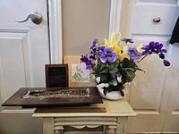 Photo showing floral arrangement, tealight holder, small framed photo, and wall art on a small white table against a door.