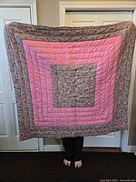 Front view of the quilt held up, showing a large central pink square with surrounding decorative floral and paisley borders, held by person standing barefoot indoors.