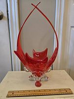Front view of tall, orange-red art glass bowl with clear glass footed base and long curved handles extending upwards.