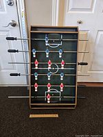 View of the entire tabletop foosball game showing wooden frame, green playing field with white markings, metal rods with blue and red player figurines, and black plastic handles. Ruler at base for scale showing about 27 inches width.