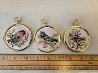 Three small needlepoint bird wall hangings arranged in a row on a beige surface with a ruler below them, showing approximate size of 3 inches diameter each, with brass-colored frames and hanging loops.