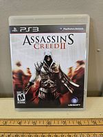 Front of PS3 game case showing Assassin's Creed II cover art with character and logos.