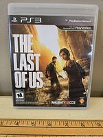Photo of PlayStation 3 game The Last of Us with cover art showing characters Joel and Ellie and post-apocalyptic city background.