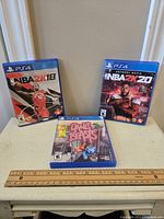 Three PS4 game cases: NBA 2K18, NBA 2K20, and Gang Beasts displayed on a small table with a ruler for scale