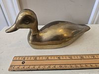 Solid brass duck figurine placed on a white surface with a ruler showing approximate length of 7 inches