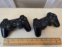 Two black Sony PS3 DualShock 3 wireless controllers side by side on a wooden surface with a ruler indicating size.